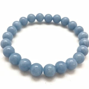 Könnte beinhalten: Ein hellblaues Perlenarmband. Das Armband besteht aus runden, glatten Perlen, die zu einem Kreis aufgefädelt sind. Die Perlen haben eine weiche, gedämpfte blaue Farbe, und das Armband liegt vor einem weißen Hintergrund.