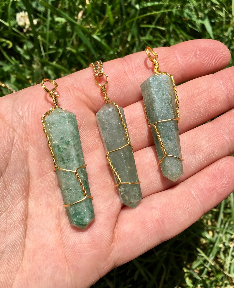 Puede incluir: Tres colgantes de cristal hexagonal verde, semitransl&uacute;cido, cada uno envuelto en un fino alambre de color dorado. Los cristales presentan inclusiones naturales y variaciones de color, con un lazo de alambre dorado en la parte superior.