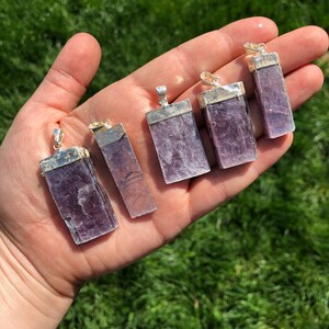 May include: Six purple lepidolite crystal pendants with silver bail settings. The pendants are rectangular in shape and have a natural, raw finish.