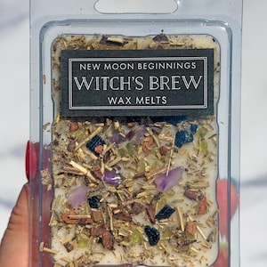May include: A rectangular plastic clamshell container holding off-white wax melts topped with dried herbs, small purple amethyst stones, and dark blue mineral fragments. A black label reads NEW MOON BEGINNINGS WITCH'S BREW WAX MELTS in white text.