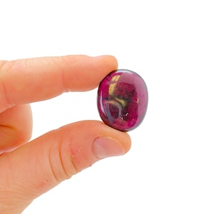 May include: A close-up of a small, oval-shaped, dark purple gemstone with gold flecks. The gemstone is being held between two fingers.