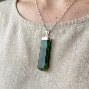 May include: A silver necklace featuring a dark green, rectangular jade pendant. The jade has a polished, faceted design and is suspended from a delicate silver chain. The pendant is approximately 2 inches long.
