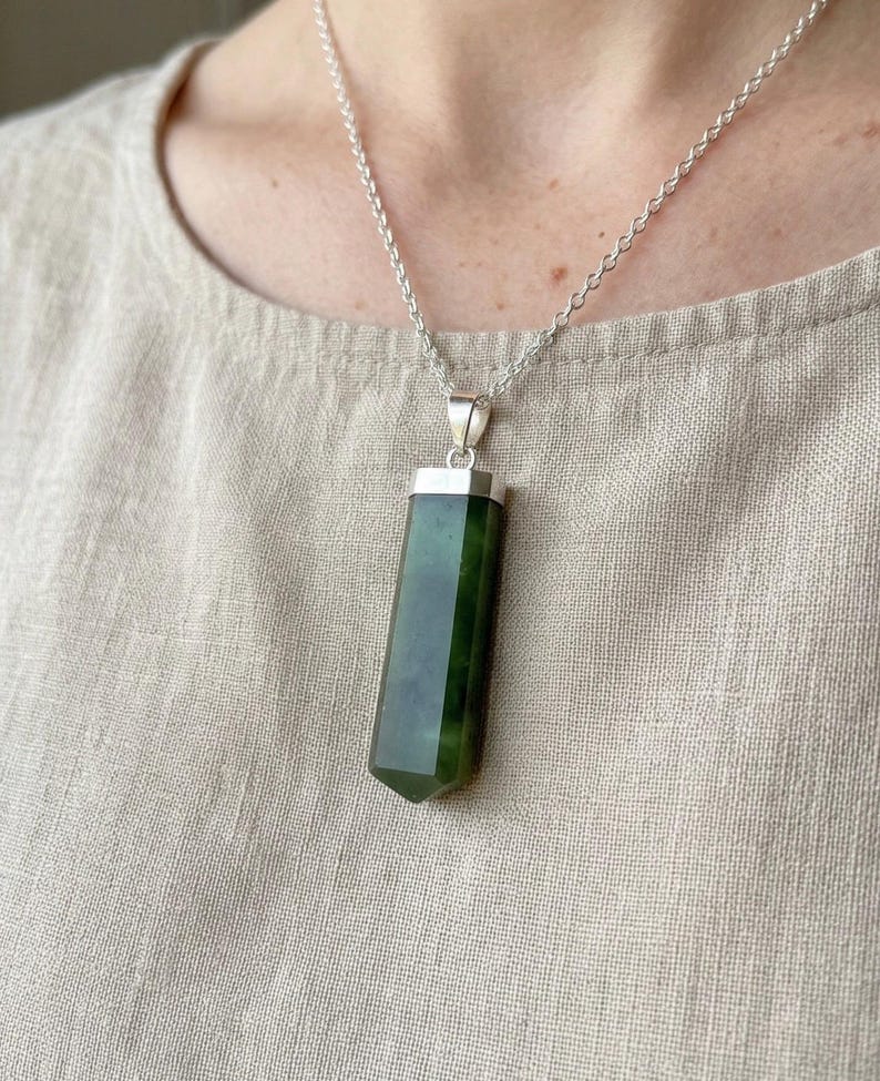 May include: A silver necklace featuring a dark green, rectangular jade pendant. The jade has a polished, faceted design and is suspended from a delicate silver chain. The pendant is approximately 2 inches long.