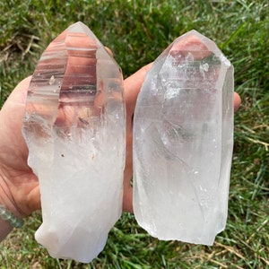 May include: Two clear quartz crystal points, one slightly larger than the other, with a natural, raw finish. The crystals are held in a hand against a green grassy background.