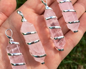 May include: Four pink rose quartz crystal pendants wrapped in silver wire. The pendants are of varying sizes and are displayed on a hand.