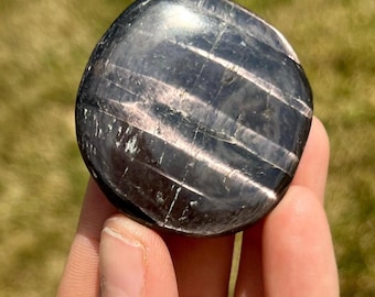 Puede incluir: Una piedra preciosa lisa, negra y plateada con un patrón estriado. La piedra preciosa está pulida y tiene una superficie reflectante.