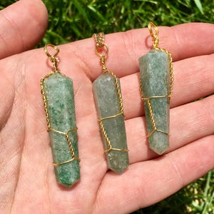 Puede incluir: Tres colgantes de cristal de aventurina verde, cada uno con forma de punta hexagonal y envuelto en un fino alambre de color dorado. Cada colgante presenta un peque&ntilde;o lazo de alambre en la parte superior, mostrando la textura natural moteada.
