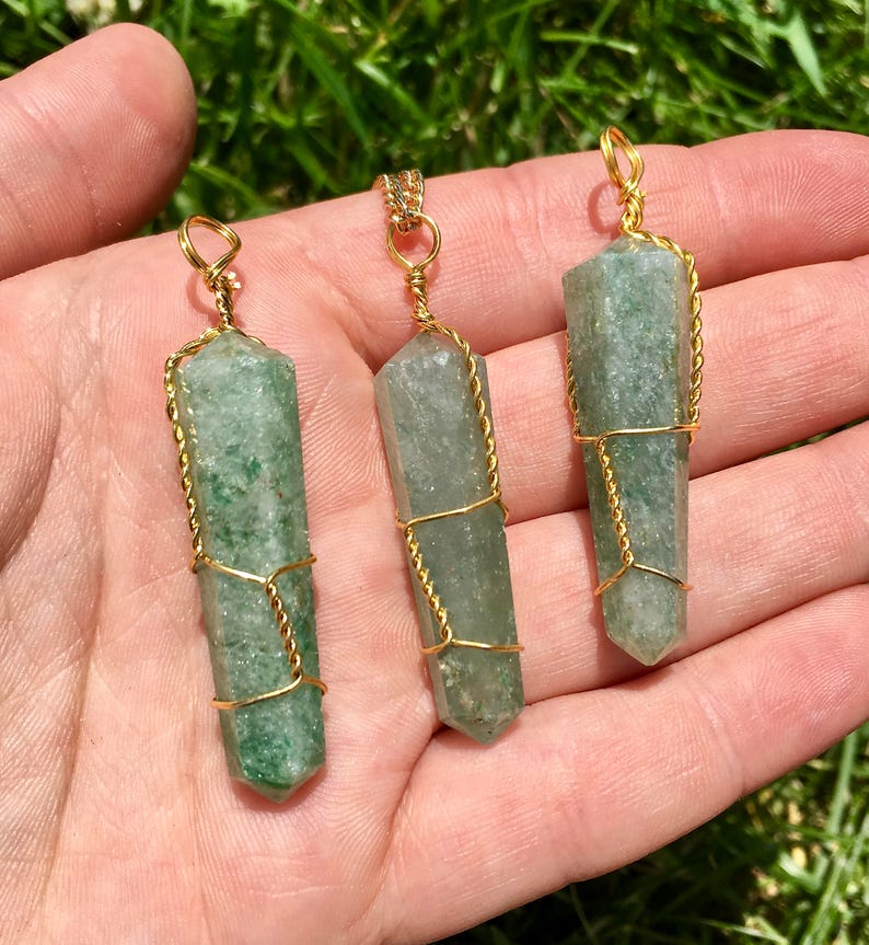 Puede incluir: Tres colgantes de cristal de aventurina verde, cada uno con forma de punta hexagonal y envuelto en un fino alambre de color dorado. Cada colgante presenta un peque&ntilde;o lazo de alambre en la parte superior, mostrando la textura natural moteada.