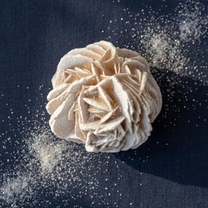 May include: A desert rose crystal formation with layered, petal-like structures in a pale beige color. The mineral specimen features sharp, crystalline edges and a dense, clustered arrangement, surrounded by scattered fine silver glitter.