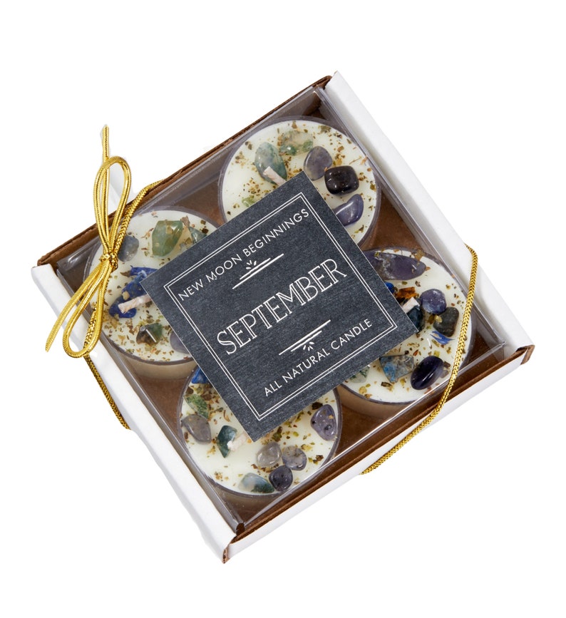 K&ouml;nnte beinhalten: Ein Set mit vier wei&szlig;en Teelichtern mit eingebetteten Kristallen und goldenem Glitzer. Die Kerzen sind in einer wei&szlig;en Schachtel mit einem durchsichtigen Deckel und einem schwarzen Etikett verpackt, auf dem "New Moon Beginnings September All Natural Candle" steht.