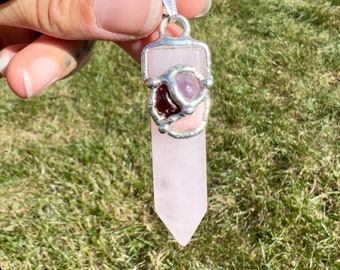 Op de afbeelding: Een zilveren hanger met een roze rozenkwartspunt en een zilveren bezel met een roze en paarse steen. De hanger is aan een zwart koord.