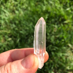 May include: A clear quartz crystal point with a natural, raw finish. The crystal is held in a hand against a blurred background of green grass.