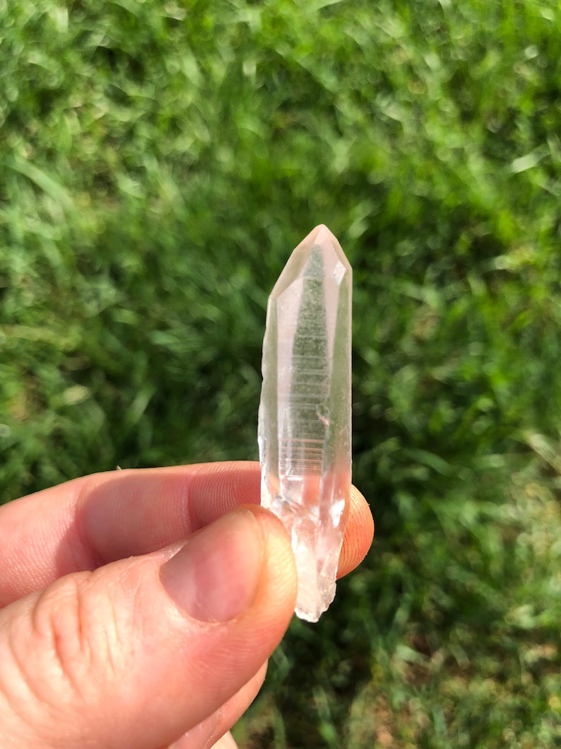 May include: A clear quartz crystal point with a natural, raw finish. The crystal is held in a hand against a blurred background of green grass.
