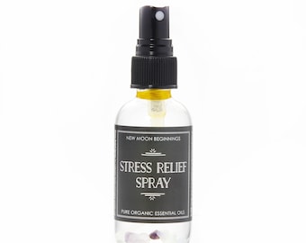 May include: A clear glass spray bottle with a black pump and a black label. The label reads "STRESS RELIEF SPRAY" and "PURE ORGANIC ESSENTIAL OILS". The bottle contains small, light purple crystals. The top of the bottle reads "NEW MOON BEGINNINGS".