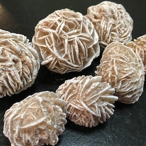May include: A collection of desert rose crystals, showcasing a cluster of beige and off-white formations. The crystals have a layered, petal-like appearance, with a rough, textured surface. The overall aesthetic is natural and earthy.