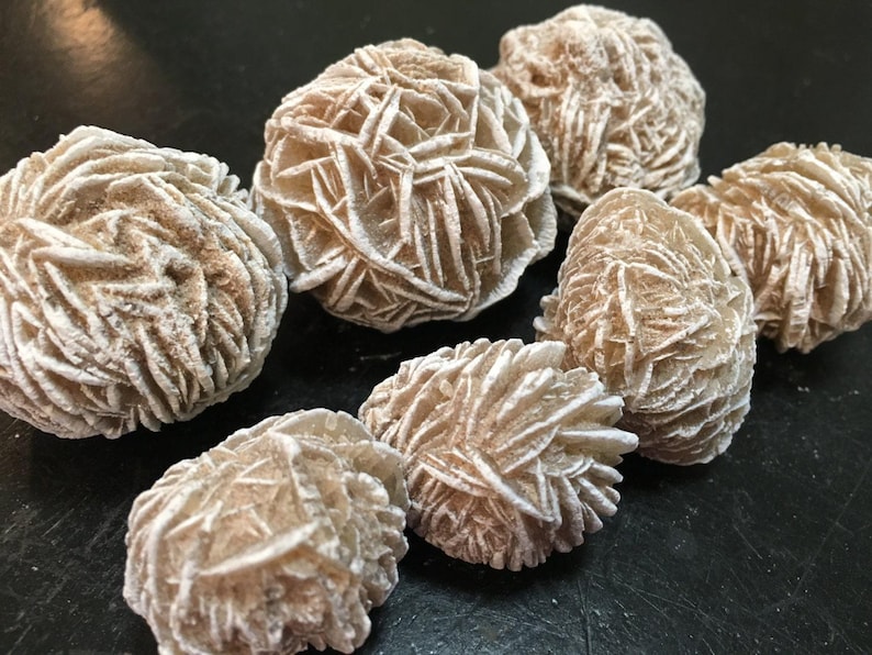 May include: A collection of desert rose crystals, showcasing a cluster of beige and off-white formations. The crystals have a layered, petal-like appearance, with a rough, textured surface. The overall aesthetic is natural and earthy.