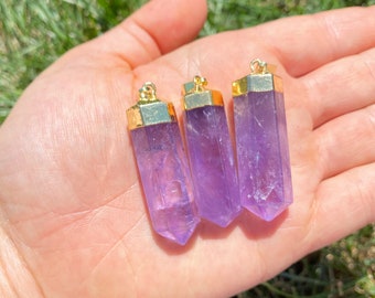 Könnte beinhalten: Drei Amethyst-Kristallanhänger mit goldfarbenen Kappen werden in einer Hand gehalten. Die Kristalle sind von transluzenter violetter Farbe und sind sechseckig geschnitten. Die Anhänger werden vor einem Hintergrund aus grünem Gras präsentiert.