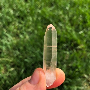 May include: A clear quartz crystal with a pink hue, pointed at both ends, held in a hand against a blurred green background.
