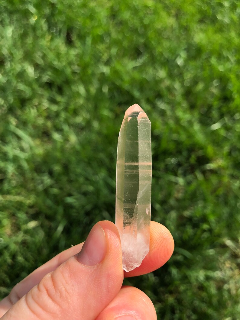 May include: A clear quartz crystal with a pink hue, pointed at both ends, held in a hand against a blurred green background.