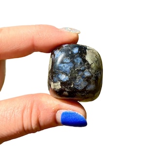 May include: A polished, square-shaped stone with a dark base color, speckled with blue and gray patterns. The stone is held between two fingers, showcasing its smooth, rounded edges and natural variations in color and texture.