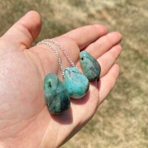 Puede incluir: Un collar de cadena de plata con tres colgantes de piedra preciosa verde esmeralda. Las piedras preciosas son lisas y tienen una forma natural irregular.