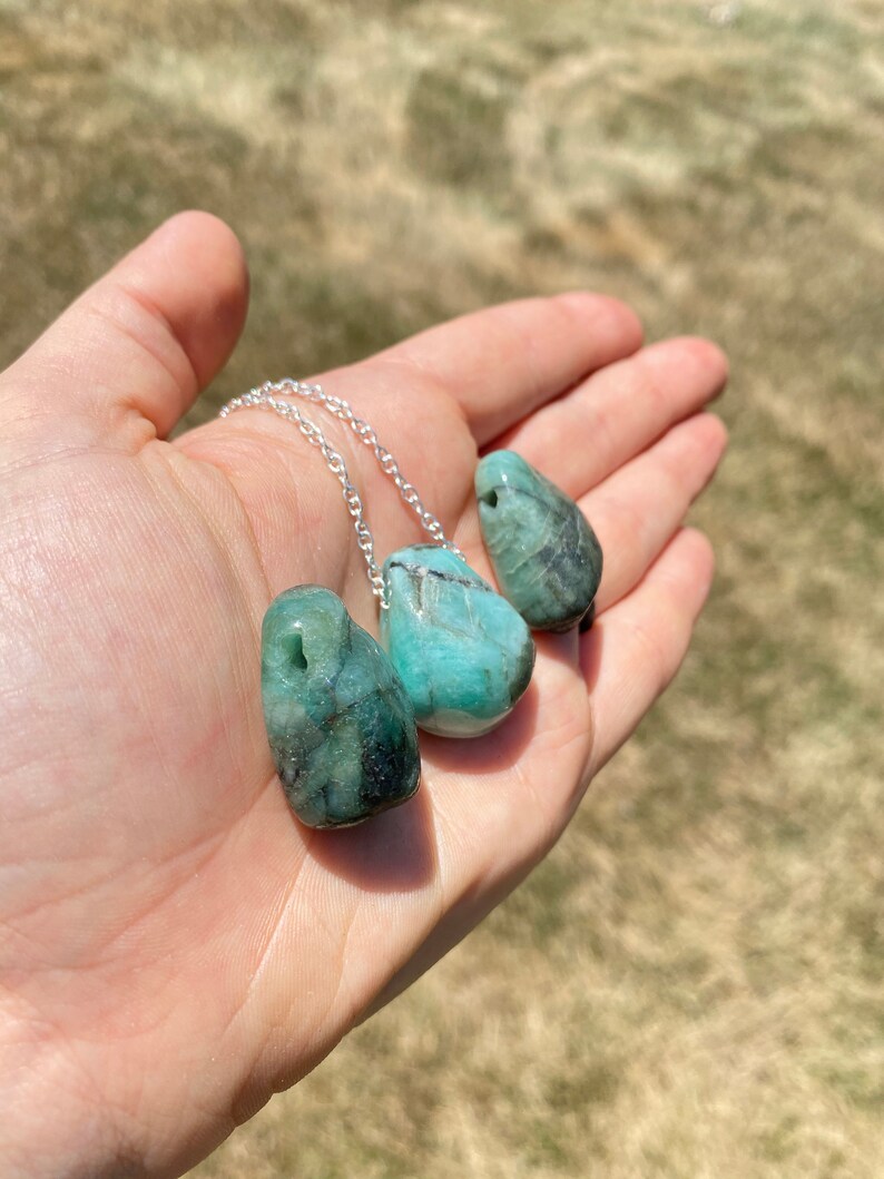 Puede incluir: Un collar de cadena de plata con tres colgantes de piedra preciosa verde esmeralda. Las piedras preciosas son lisas y tienen una forma natural irregular.