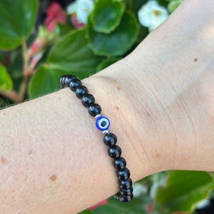 May include: Black beaded bracelet with a single blue and white evil eye charm. The bracelet is made of small, round, black beads. The evil eye charm is a bright blue with a white center and a silver-colored metal setting.