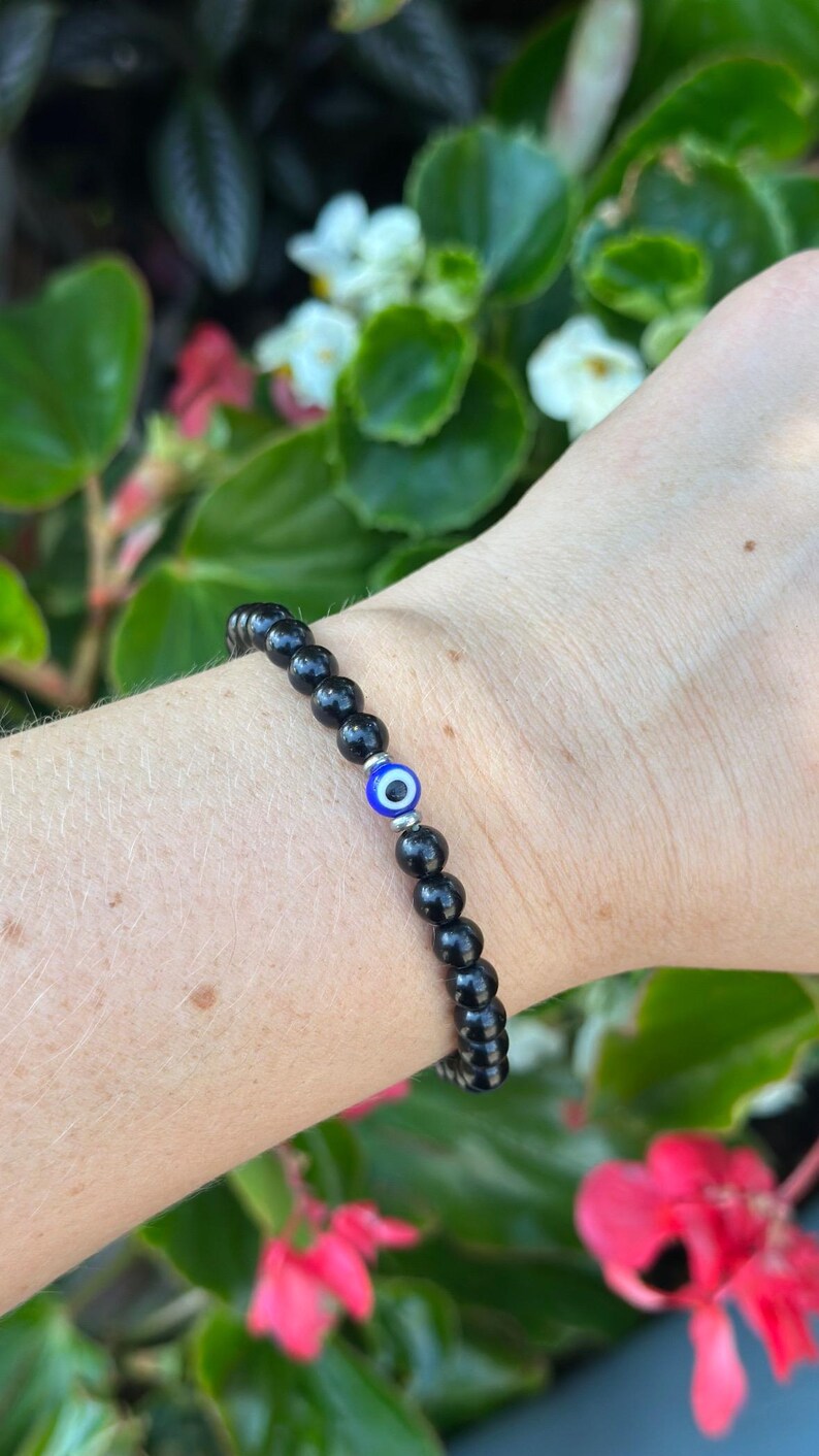 May include: Black beaded bracelet with a single blue and white evil eye charm. The bracelet is made of small, round, black beads. The evil eye charm is a bright blue with a white center and a silver-colored metal setting.