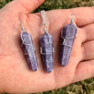 May include: Three purple lepidolite crystal pendants wrapped in silver wire. The pendants are on a hand, and one is on a silver chain.