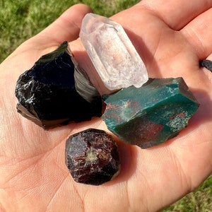 May include: A collection of raw gemstones, including a clear quartz crystal, a piece of black obsidian, a dark red garnet, and a green stone with red spots. The stones are held in an open hand against a background of green grass.