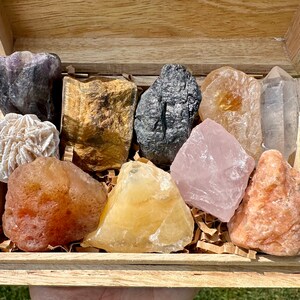 May include: A wooden box filled with a variety of colorful crystals and stones, including amethyst, tiger's eye, black obsidian, rose quartz, clear quartz, selenite, and carnelian.