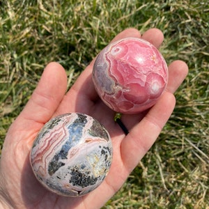 May include: Two pink and grey gemstone spheres, one with a more pronounced pink color and the other with more grey and black markings, are held in a hand.