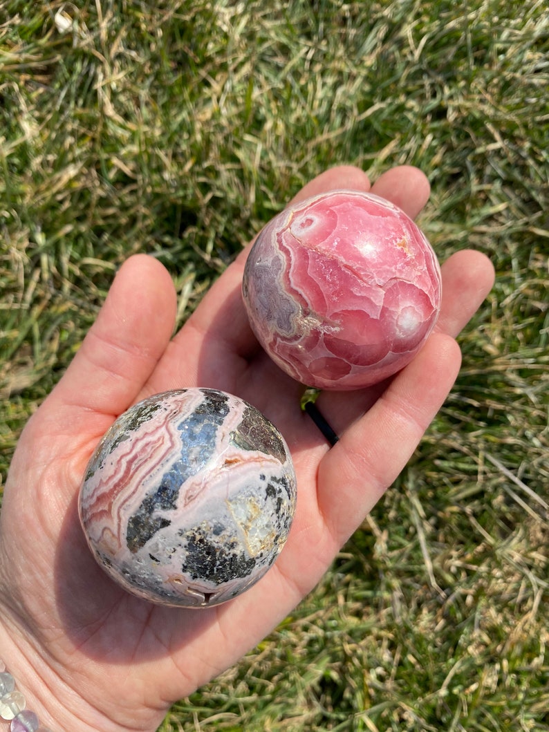 May include: Two pink and grey gemstone spheres, one with a more pronounced pink color and the other with more grey and black markings, are held in a hand.