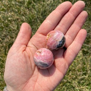 May include: Two pink and black, round, polished, gemstone spheres. The spheres are smooth and have a swirling pattern.