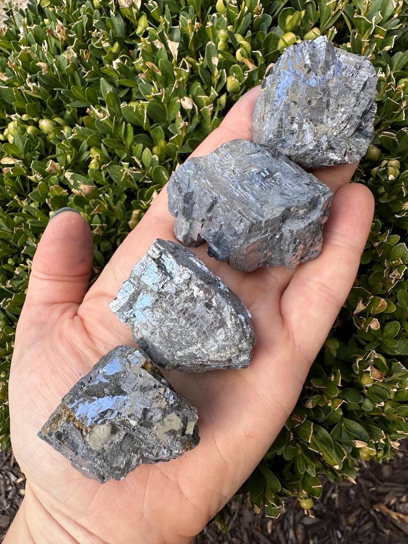 May include: Four raw, gray, metallic, crystal specimens with a silvery sheen. The crystals are rough and have a layered, striated texture.