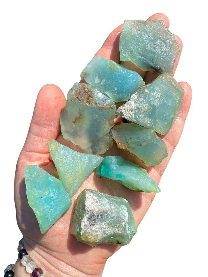 Puede incluir: Un pu&ntilde;ado de piedras preciosas en bruto de color aguamarina. Las piedras var&iacute;an en tama&ntilde;o y forma, con una calidad transl&uacute;cida. Algunas piezas tienen toques de marr&oacute;n y blanco. Las piedras preciosas se sostienen en una mano abierta.