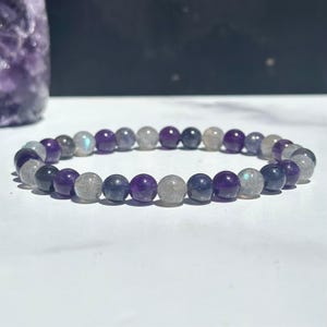 May include: A beaded bracelet featuring alternating round polished beads in deep purple amethyst and iridescent grey labradorite. The stones are arranged in a repeating pattern, showcasing the natural color variations and light-reflecting properties of the minerals.