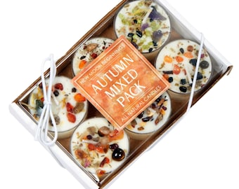 May include: A rectangular white box containing six round candles topped with various colorful stones and dried herbs. A square orange label in the center reads NEW MOON BEGINNINGS AUTUMN MIXED PACK ALL NATURAL CANDLES. The box is tied with white string.