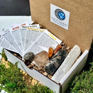May include: An open cardboard box filled with various crystals and informational cards. The crystals include clear quartz, citrine, and selenite. The box has a sticker that says "NEW MOON BEGINNINGS". The box is surrounded by green moss.