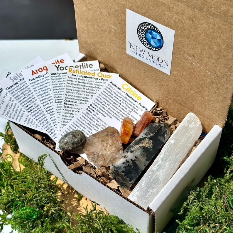 May include: An open cardboard box filled with various crystals and informational cards. The crystals include clear quartz, citrine, and selenite. The box has a sticker that says "NEW MOON BEGINNINGS". The box is surrounded by green moss.