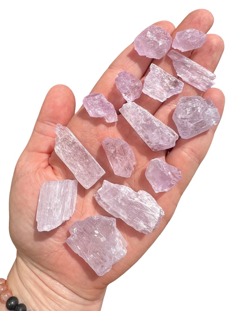 Puede incluir: Una colecci&oacute;n de cristales de color p&uacute;rpura claro de varias formas y tama&ntilde;os, sostenidos en una mano abierta. Los cristales tienen una apariencia transl&uacute;cida y una textura ligeramente rugosa, con estructuras internas visibles. El fondo es blanco liso.