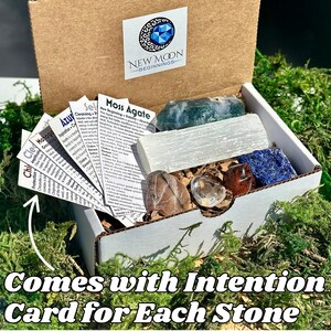 May include: A gift box labeled "New Moon Beginnings" filled with various crystals and intention cards. The box contains a variety of stones, including a large selenite wand, moss agate, and a blue lapis lazuli. The text "Comes with Intention Card for Each Stone" is visible.