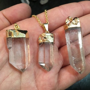 Puede incluir: Tres colgantes de cristal de cuarzo transparente con tapas chapadas en oro. Los colgantes est&aacute;n en una cadena de oro y se sostienen en una mano.