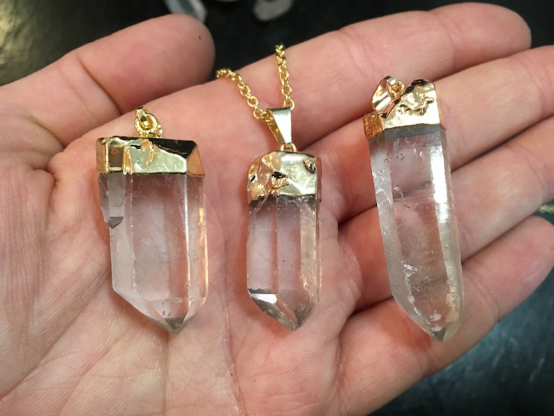 Puede incluir: Tres colgantes de cristal de cuarzo transparente con tapas chapadas en oro. Los colgantes est&aacute;n en una cadena de oro y se sostienen en una mano.