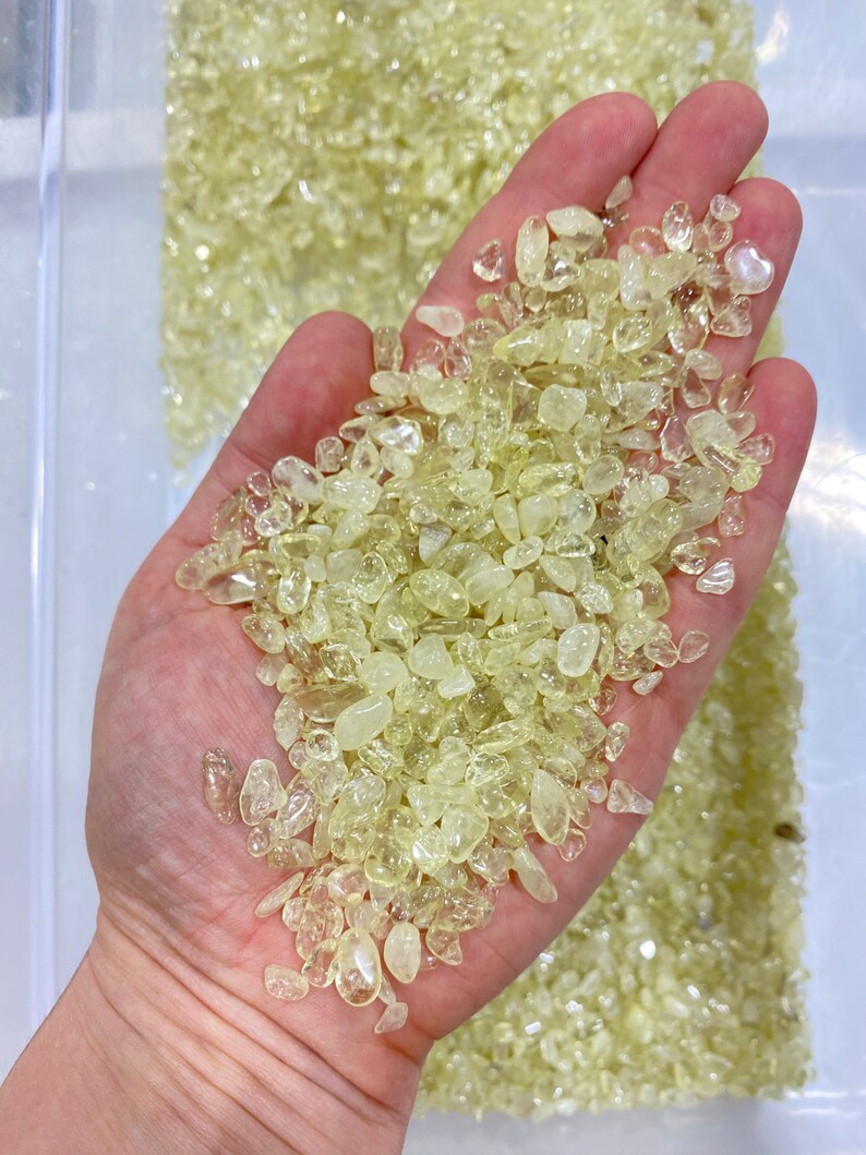 May include: A hand holding a pile of small, translucent, light yellow citrine crystals. The crystals are tumbled and have a smooth, polished finish.