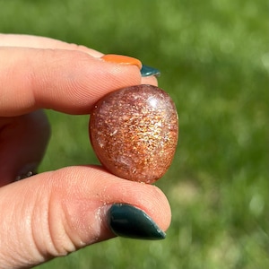 May include: A smooth, polished sunstone with a reddish-orange hue and a shimmery, iridescent finish. The stone is a natural crystal and has a unique, organic shape.