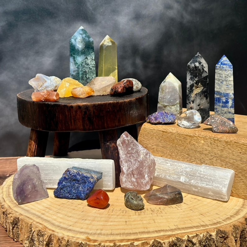 May include: A collection of various crystals and gemstones, including towers and raw stones. The crystals are displayed on a small wooden stool and a wooden slab. Colors include green, yellow, blue, purple, and clear.