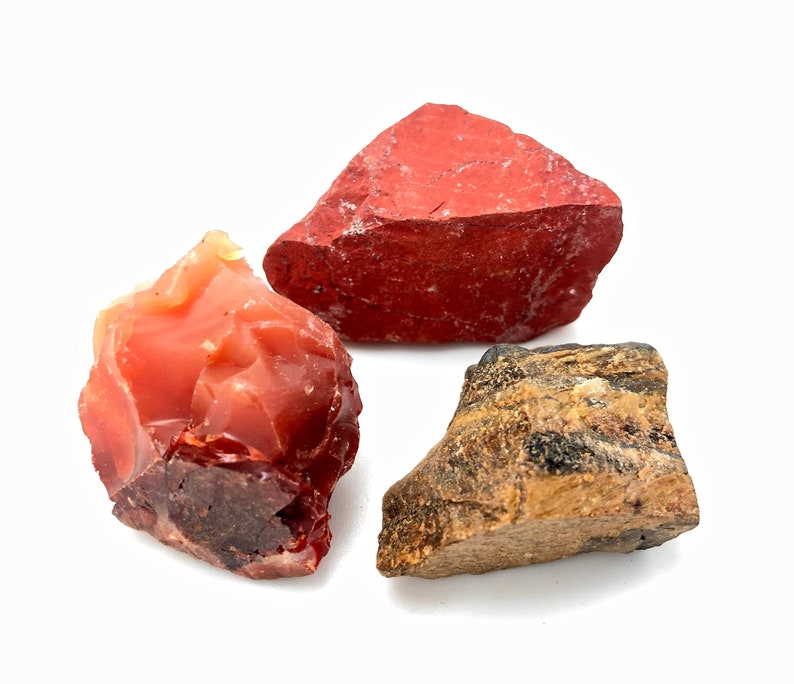May include: Three rough, natural stones: a red jasper, a carnelian, and a tiger's eye. The stones are all different shapes and sizes, and they have a natural, unpolished look.