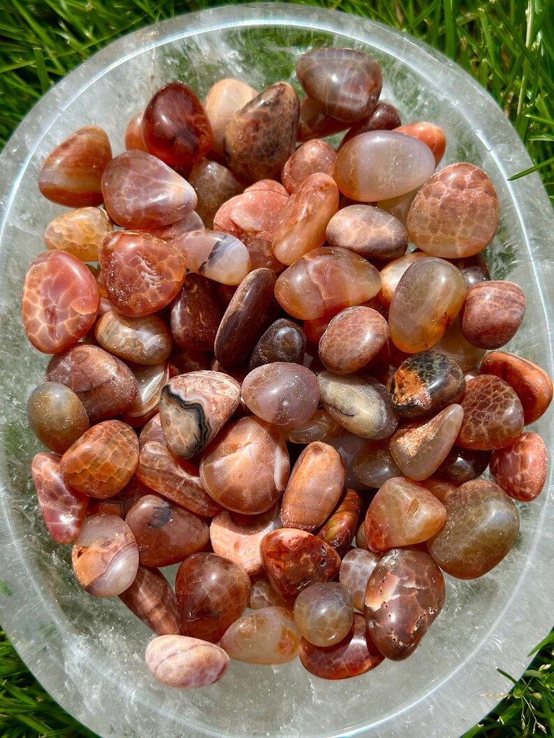 May include: A clear glass bowl filled with polished, tumbled stones. The stones are a mix of warm colors, including shades of orange, brown, and tan. The stones have a smooth, rounded shape and varying patterns.