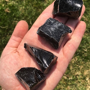 Puede incluir: Cuatro piezas de obsidiana negra en bruto con una textura brillante similar al vidrio y formas irregulares de bordes afilados. Cada piedra muestra un color negro opaco profundo con superficies reflectantes que captan la luz y muestran fracturas naturales.